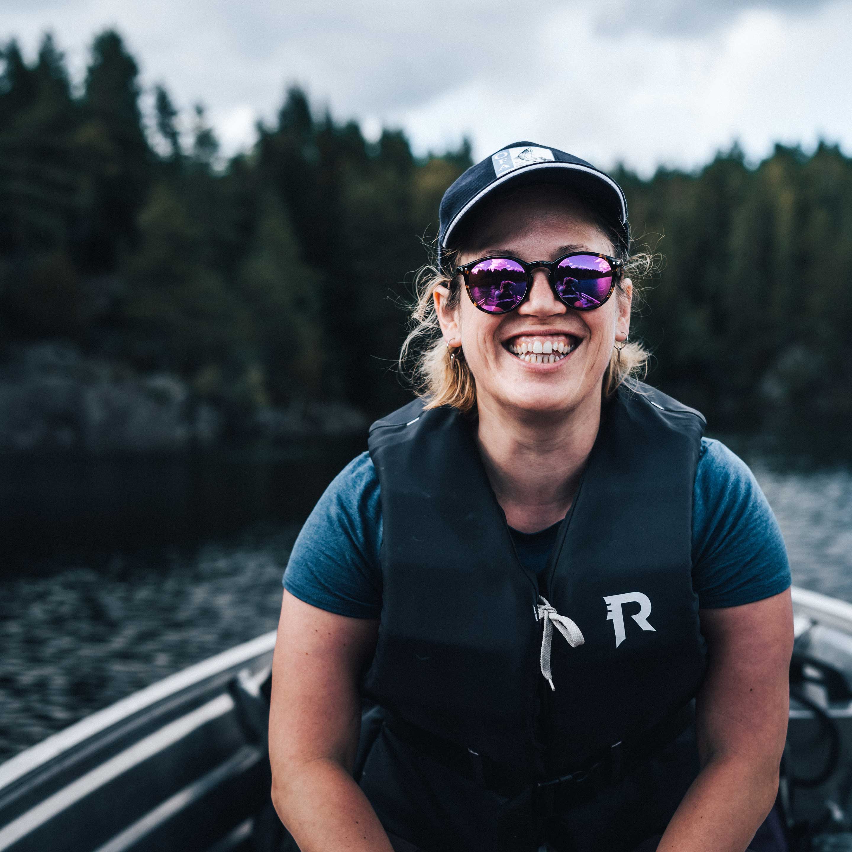 Nærbilde av smilende kvinne fra Fiskejenter i Marka, med solbriller og caps, som har på seg redningsvest og sitter i en båt.  